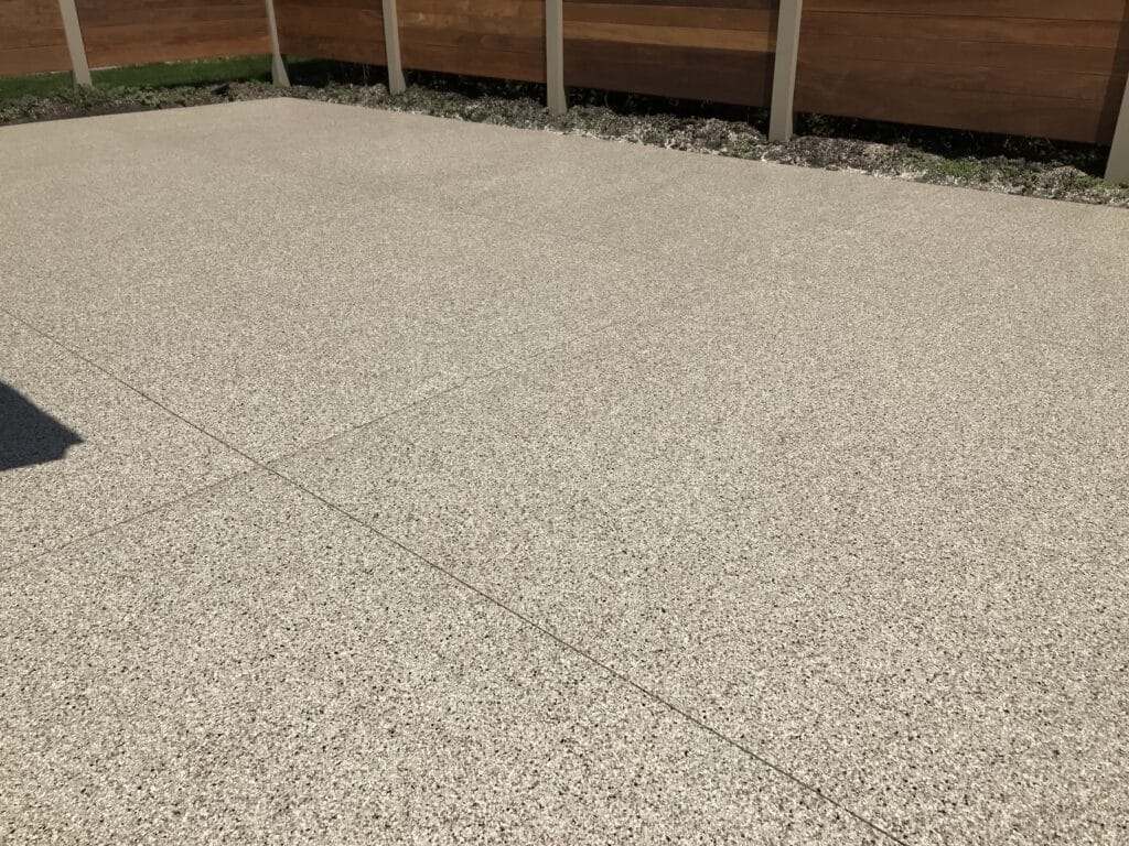 This image shows a textured concrete surface with distinct lines demarcating sections, a wooden fence in the background, and a shadow in one corner.