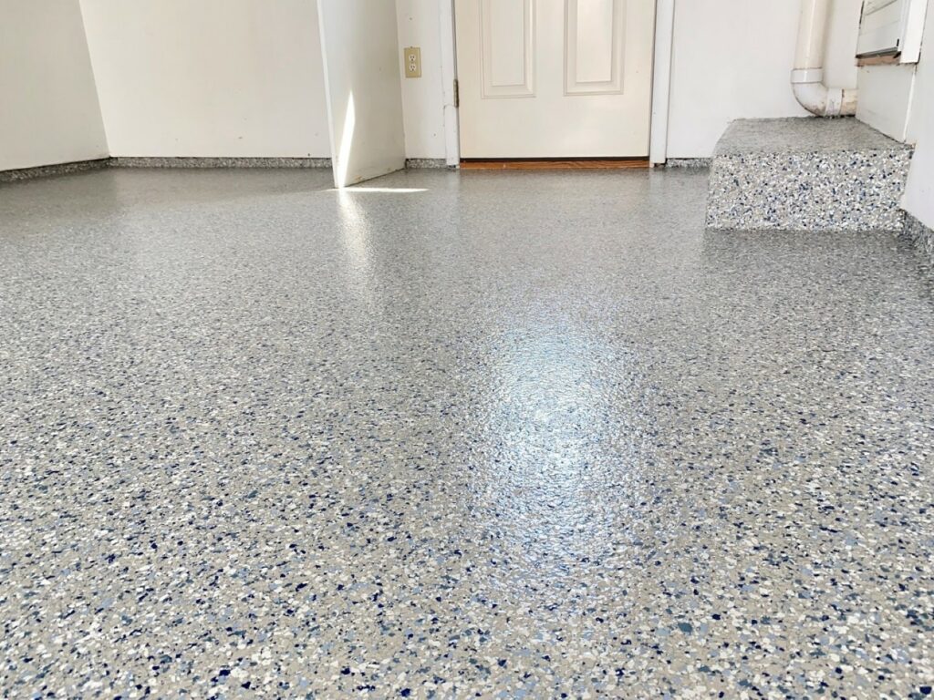 The image shows an interior space with terrazzo flooring featuring blue and white chips, white walls, a closed door, and a step with pipework alongside it.