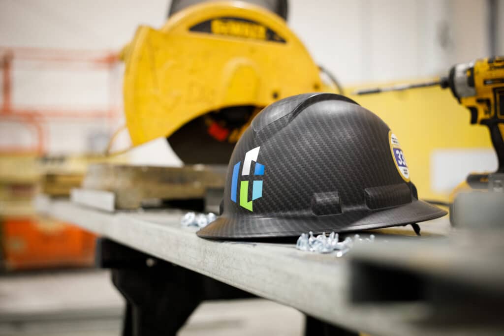 A carbon fiber patterned hard hat rests on a workbench with industrial equipment and power tools blurred in the background, symbolizing a construction environment.