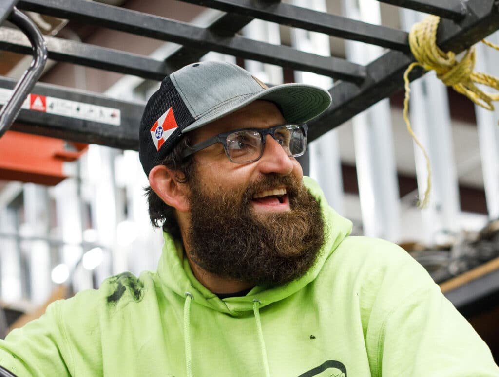 A person with a beard wears glasses, a cap, and a hi-vis hoodie, smiling beside industrial shelving with a rope hanging in the foreground.