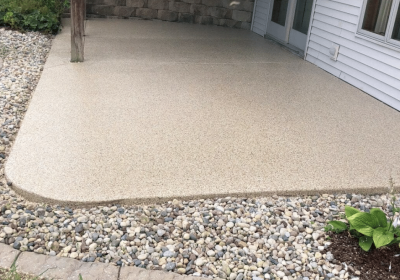 A beige concrete patio bordered by loose stones with a green plant in the corner, adjacent to a white-sided building, under a cloudy sky.